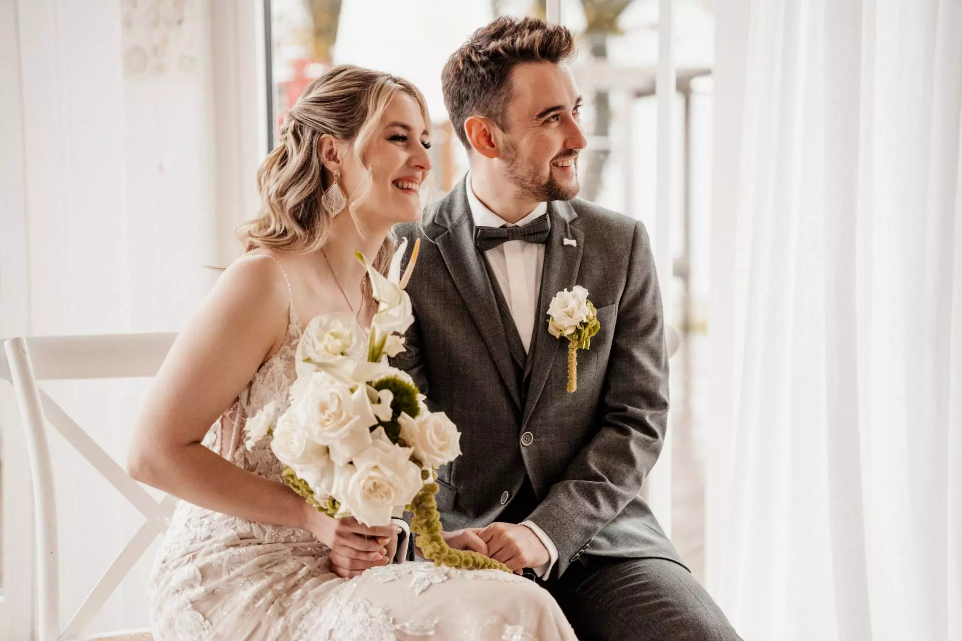 Lächelndes Brautpaar sitzt mit Blumenstrauß.