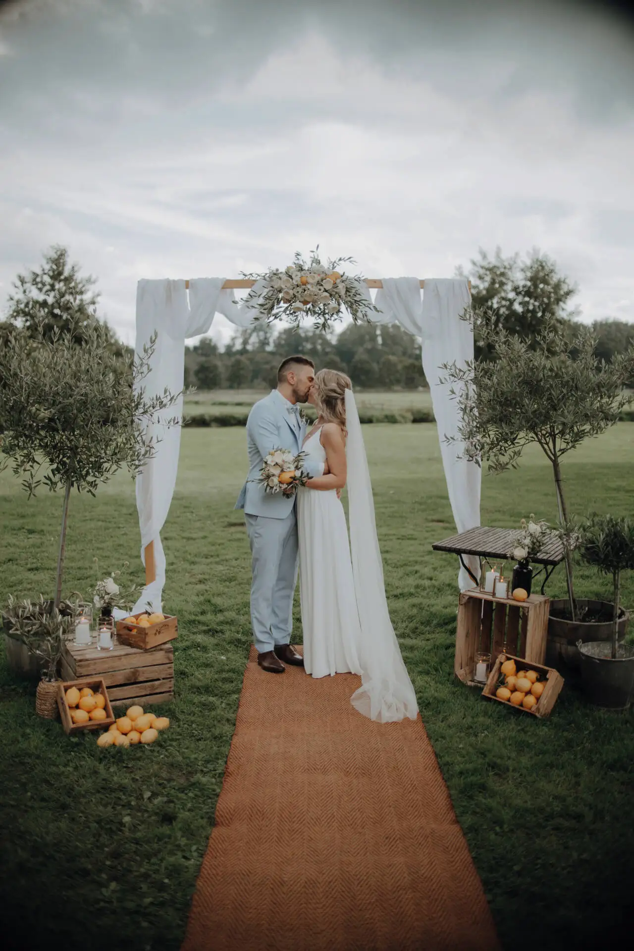 Brautpaar küsst sich unter Hochzeitsbogen im Freien.