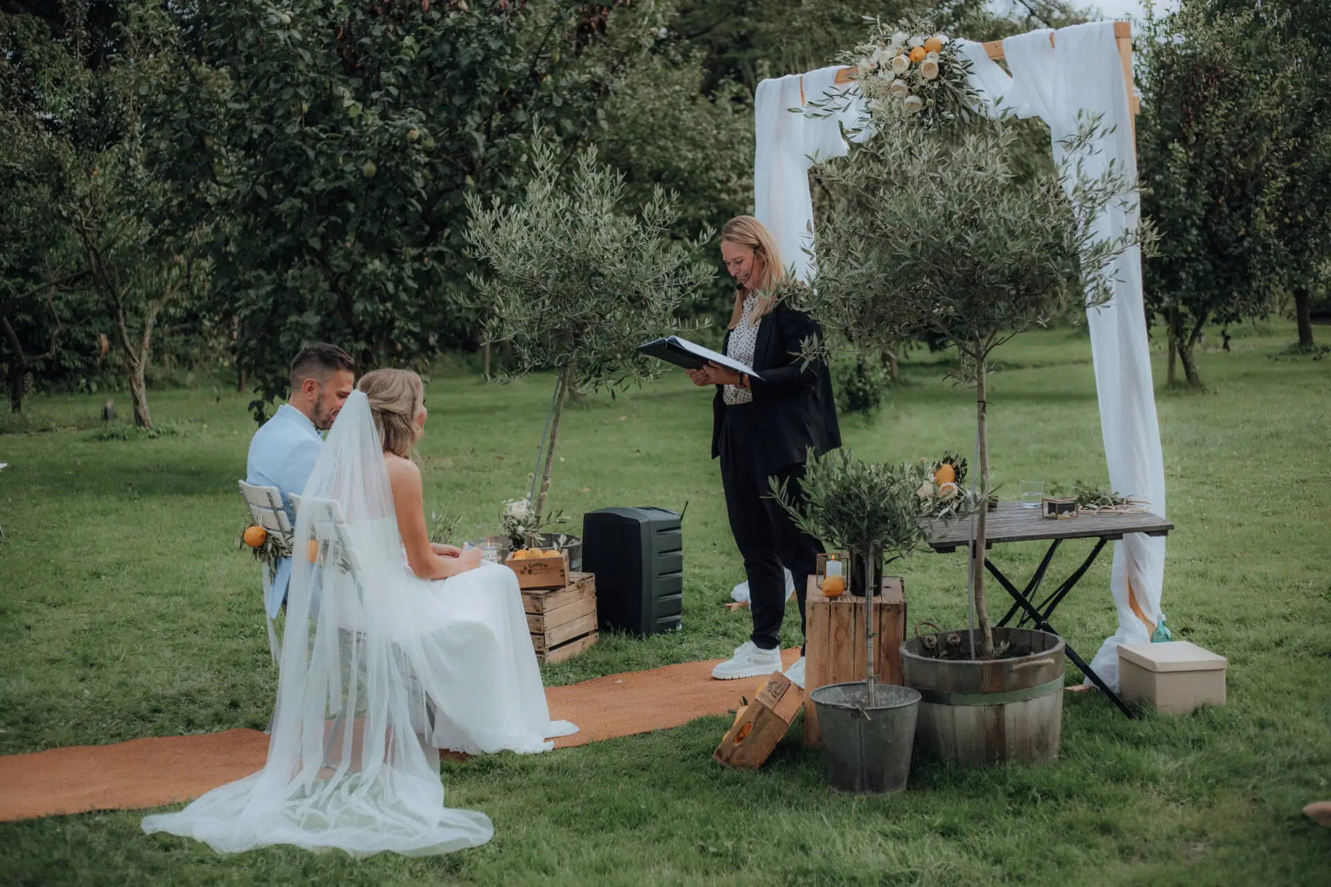 Paar heiratet draußen unter Bäumen mit Rednerin.