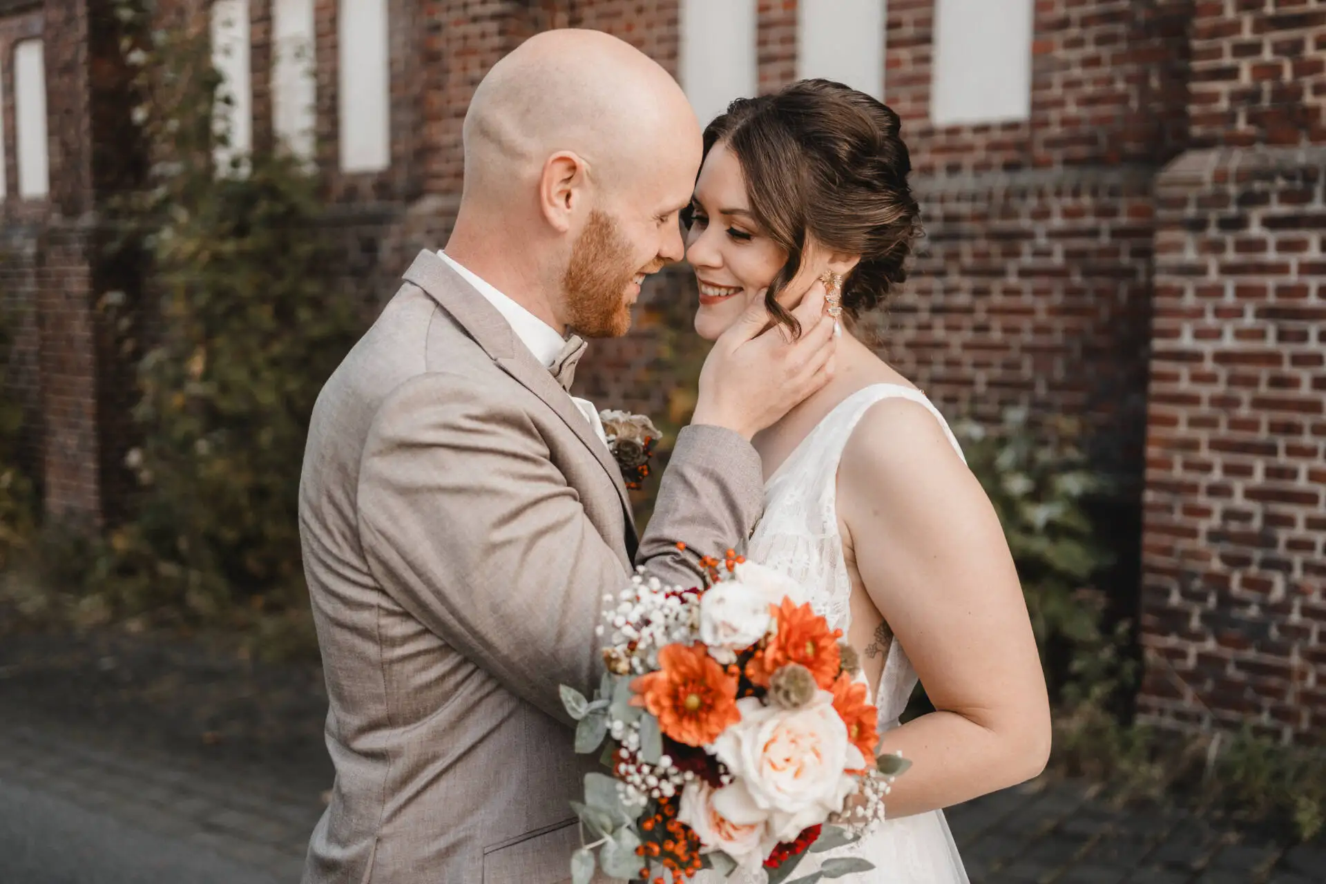 Brautpaar umarmt sich lächelnd vor Backsteinwand.