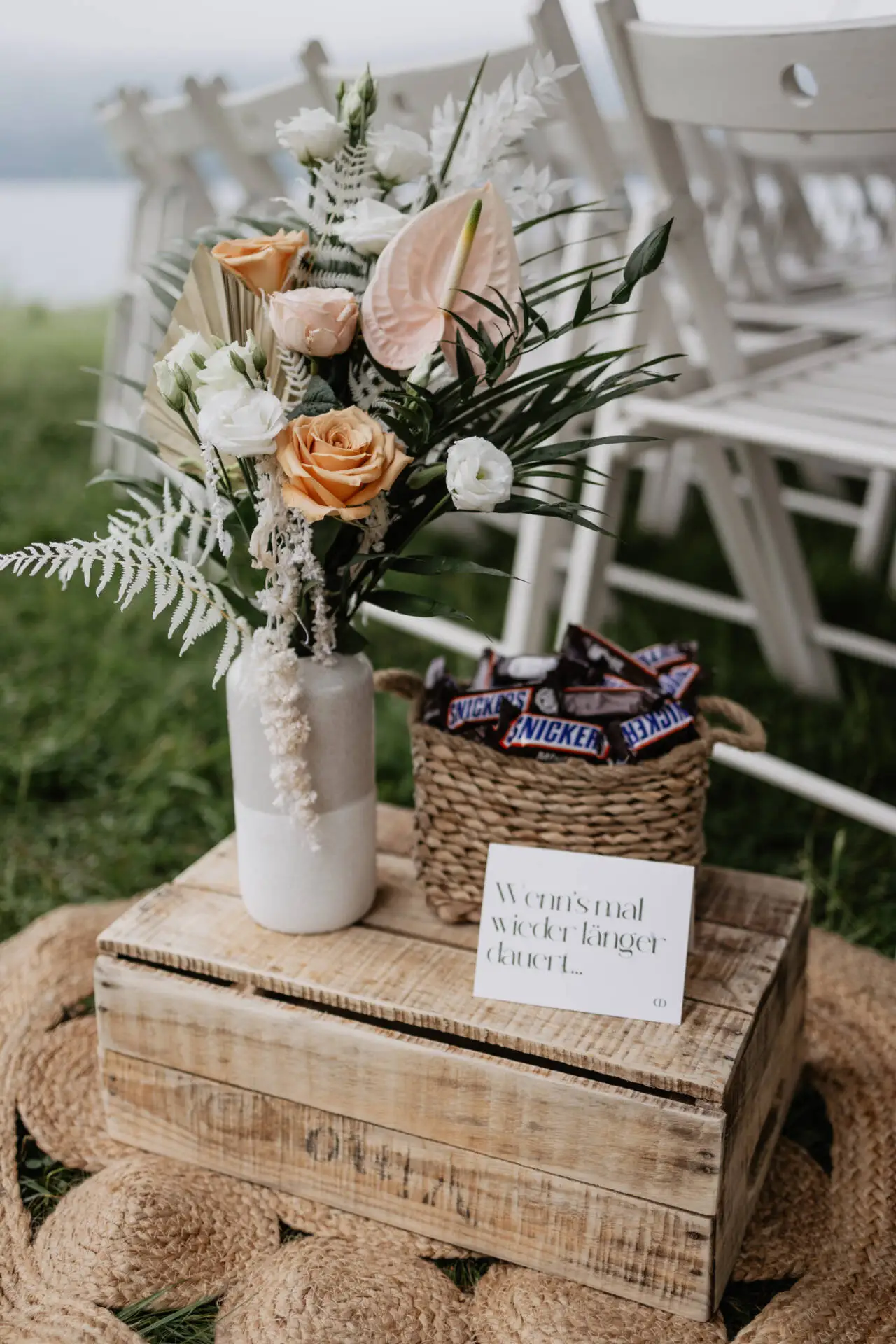 Blumenstrauß und Süßigkeitenkorb auf Holzkiste vor Stühlen.